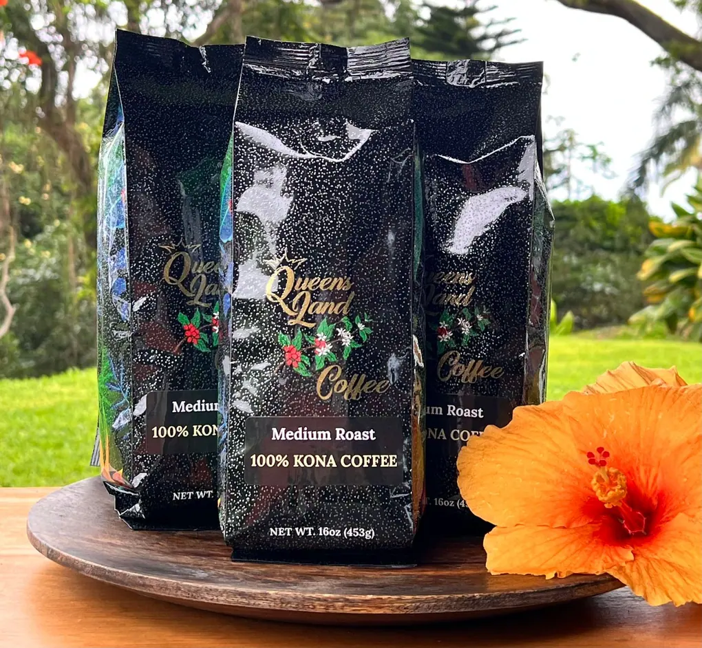 Three bags of Kona coffee, medium roast, sit on a wooden plate with an orange hibiscus flower.